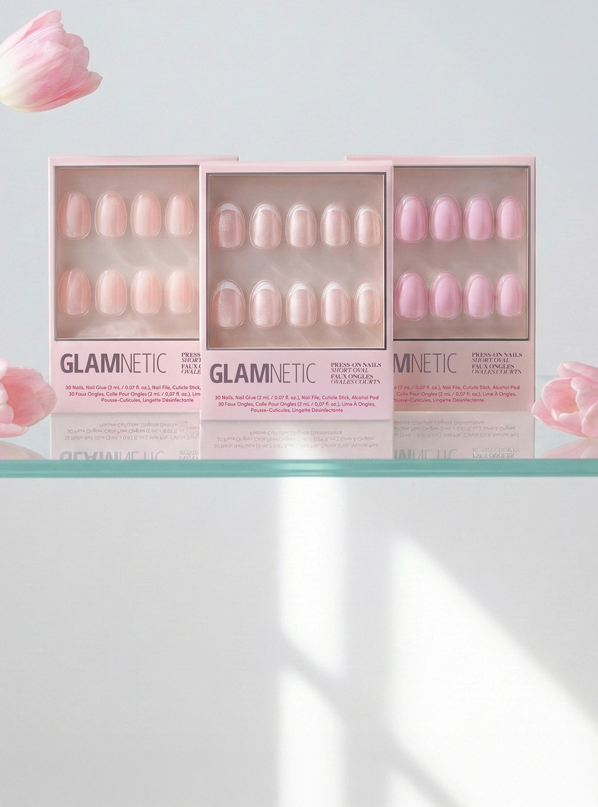 Three Glamnetic press-on nail boxes on a shelf with pink flowers in the background.